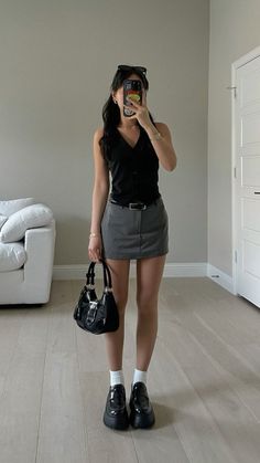 Woman in a black vest and grey mini skirt holding a black handbag indoors