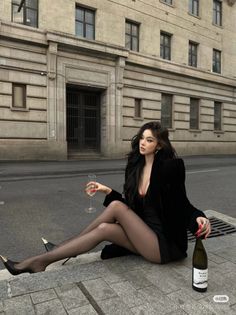 Woman in a black dress and blazer sitting on a city street with wine