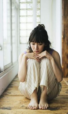Woman with dark hair and bangs sitting barefoot on a wooden floor indoors