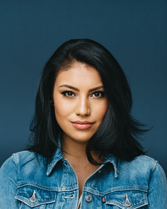 Woman with black hair wearing a denim jacket against a dark background