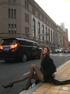 Woman in a black blazer and sheer stockings sitting on a city curb