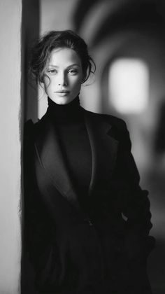 Woman in a black turtleneck and coat leaning against a wall in black and white