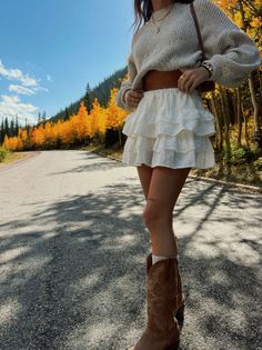 Woman in a cropped sweater, ruffle skirt, and cowboy boots on a road with autumn trees