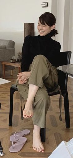 Woman in a black turtleneck and olive pants, sitting and smiling with bare feet