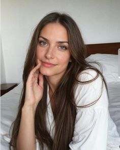 Woman with long brown hair in a white shirt, sitting on a bed