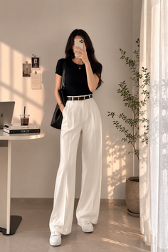 Woman in a black t-shirt and white pants taking a mirror selfie indoors