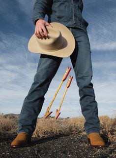 Person in denim holding a cowboy hat, standing over two large arrows in a desert landscape