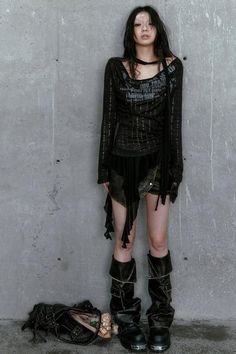 Woman in dark, distressed clothing and boots against a concrete wall