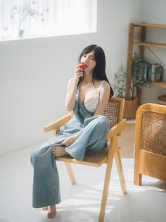 Woman in denim overalls and a white bra holding an apple while sitting on a chair