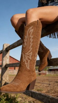 Person wearing brown cowboy boots with white embroidery, sitting on a wooden fence