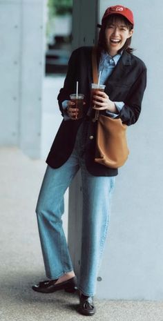 Woman in a red cap, black blazer, and jeans holding two drinks outdoors