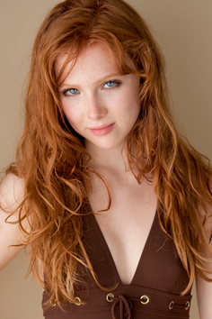 Woman with long red wavy hair and blue eyes wearing a brown top