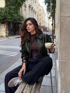 Woman in a black leather jacket and wide leg pants sitting on a bench on a city street