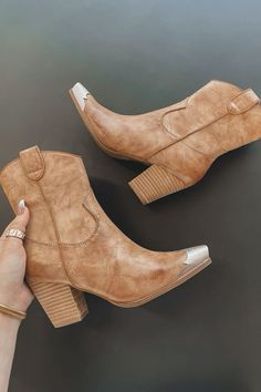 Pair of brown cowboy ankle boots with silver toe caps, held by a hand