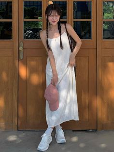 Woman in a white dress with braids holding a pink cap in front of a wooden door