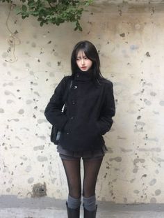 Woman with black hair wearing a black coat, shorts, and tights outdoors