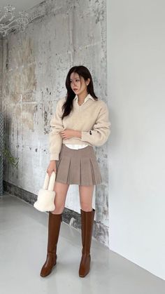 Woman in a beige sweater, pleated skirt, and brown boots holding a furry bag