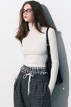 Woman with long hair wearing a white turtleneck and plaid pants, leaning against a wall