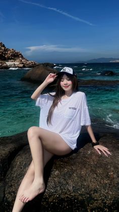 Woman in a white t-shirt and baseball cap sitting on rocks by clear blue water