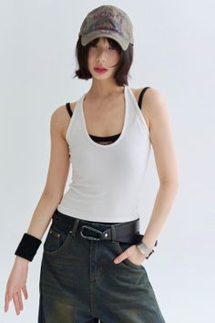 Woman with short hair and a baseball cap wearing a white tank top and jeans
