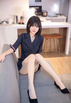 Woman with short black hair wearing a blazer dress and pantyhose, sitting on a couch