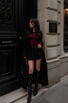 Woman in a black coat and pleated skirt standing outside a building