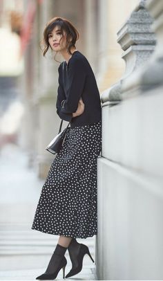 Woman in a black polka dot skirt and top leaning against a wall outdoors