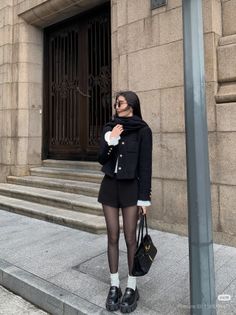 Woman in a black jacket, shorts, and tights standing on a city street