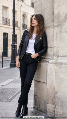 Woman in a black leather jacket and jeans leaning against a stone pillar outdoors