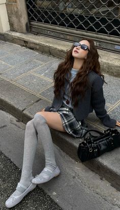 Woman with long wavy hair sitting on a curb in a plaid skirt and knee-high socks