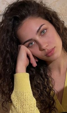 Woman with curly hair and green eyes wearing a yellow sweater, resting her head on her hand