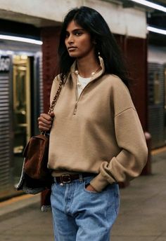 Woman with long dark hair wearing a beige sweatshirt and jeans in a subway station