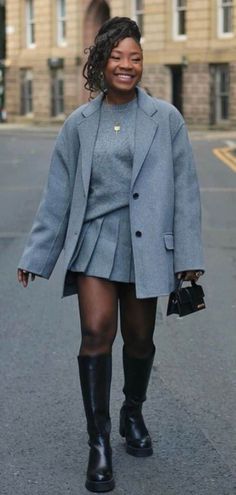 Woman in a gray blazer and pleated skirt with knee-high boots and a mini handbag