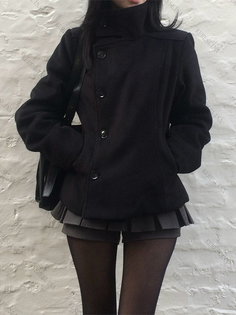 Person in a black coat, pleated skirt, and tights against a white brick wall