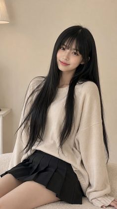 Woman with long black hair and bangs wearing a sweater and pleated skirt indoors