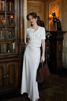 Woman in a white skirt and top with a pearl necklace holding a brown bag indoors