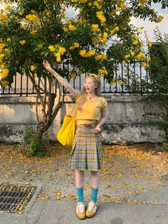 Woman in a yellow top and plaid skirt posing outdoors with yellow flowers