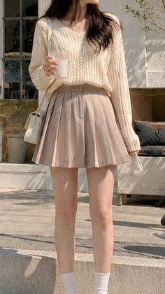 Person in a cream knitted sweater and pleated skirt holding a drink outdoors