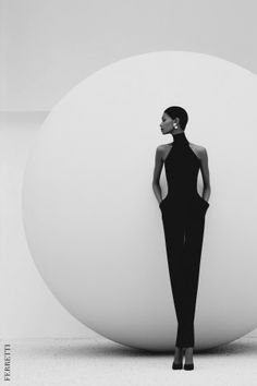 Woman in a black jumpsuit with short hair and pearl earrings against a white sphere