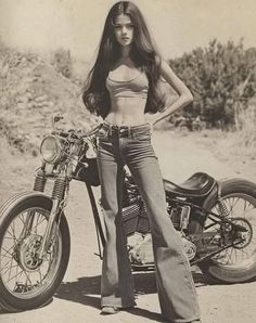 Woman in a crop top and bell bottom jeans standing next to a motorcycle