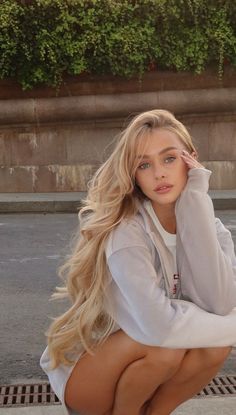 Woman with long blonde hair and blue eyes in a hoodie, crouching outdoors