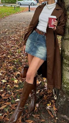 Person in a brown trench coat and denim skirt holding a coffee cup outdoors