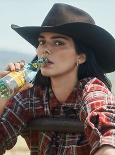 Woman in a cowboy hat and plaid shirt drinking from a bottle outdoors