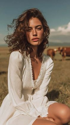 Woman with curly hair in a white dress sitting in a field with cows