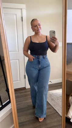 Woman in a black tank top and blue jeans taking a mirror selfie indoors