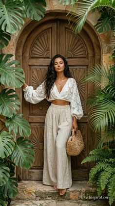 Woman in a lace top and wide leg pants holding a straw bag in front of a wooden door