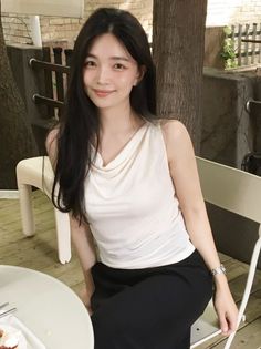 Woman with long black hair wearing a white top and black pants, sitting outdoors
