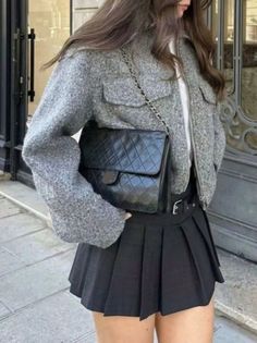 Person in a grey jacket and black pleated skirt holding a black handbag outdoors