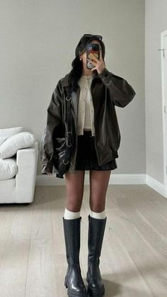 Person in a leather jacket, black skirt, and knee-high boots taking a mirror selfie