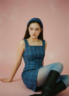 Woman in a blue plaid dress, blue tights, and black boots sitting against a pink background